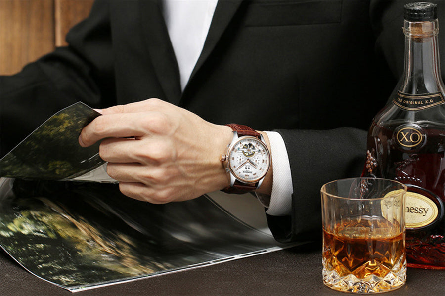 Man in a suit with a watch, glass of whiskey, and bottle of Hennessy on a table.