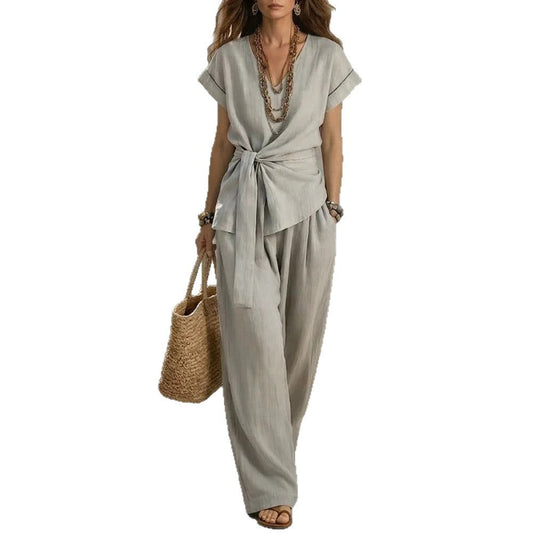 A woman modeling a light gray wrap-style short-sleeve jumpsuit with wide-leg pants, accessorized with layered necklaces, bracelets, and a woven straw tote bag against a plain white background.