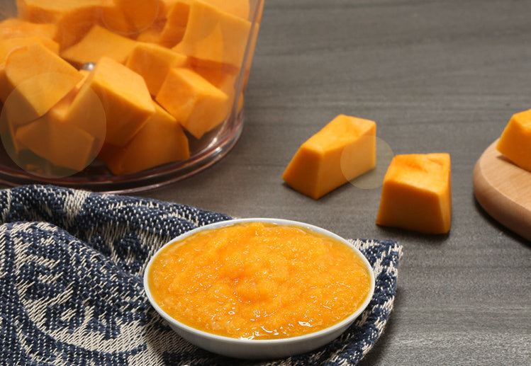 Butternut squash cubes in a glass container with a bowl of puree on a gray surface.
