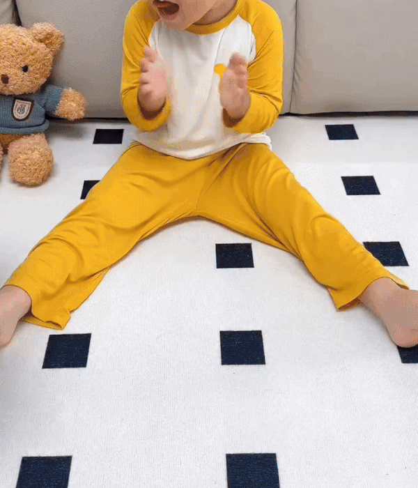 Toddler sitting on a white play mat with small dark squares and a teddy bear nearby
