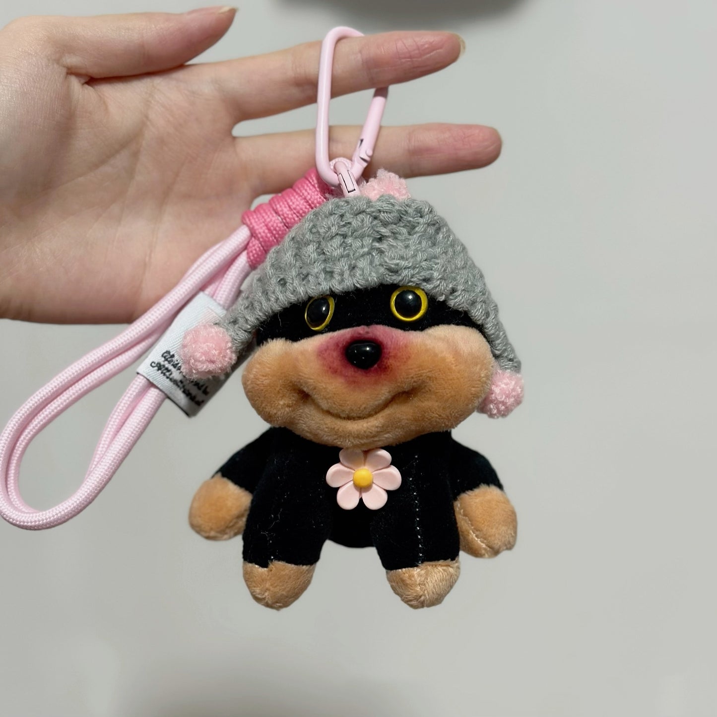 Plush keychain with a gray hat and pink accents held by a hand against a plain background