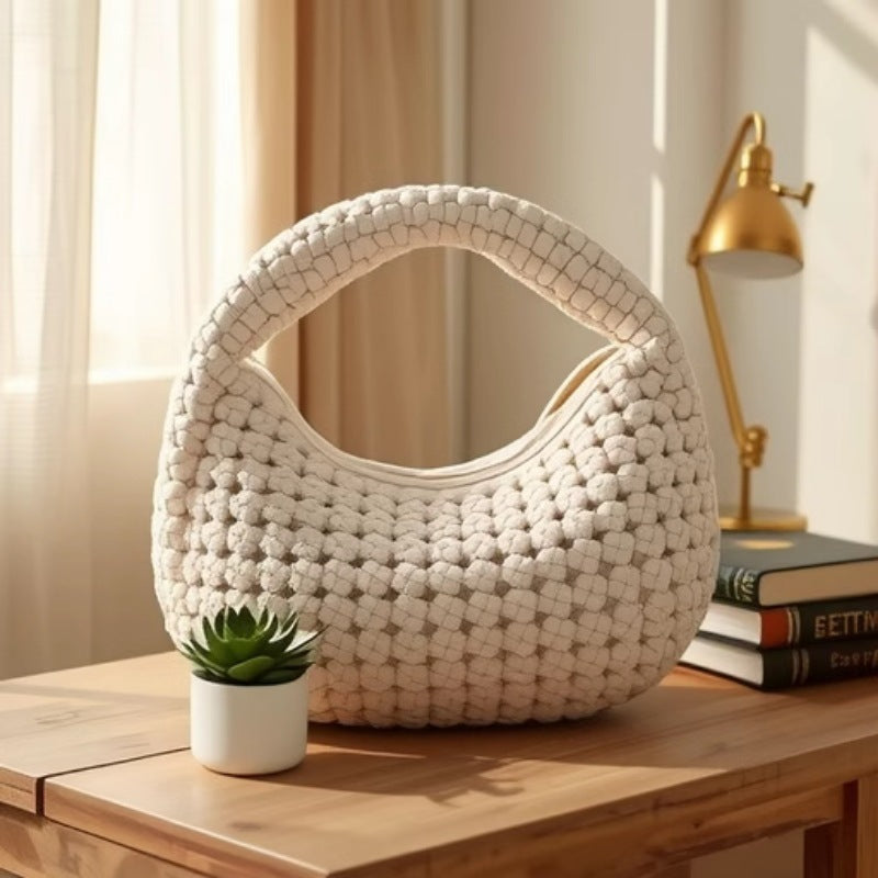 White woven handbag on a wooden surface with a plant and books in the background