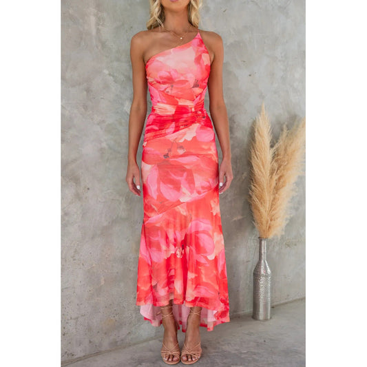 Woman wearing a pink and red one-shoulder dress against a concrete wall with a vase of dried plants.