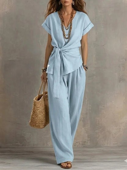 Woman wearing a light blue jumpsuit holding a straw bag against a neutral background