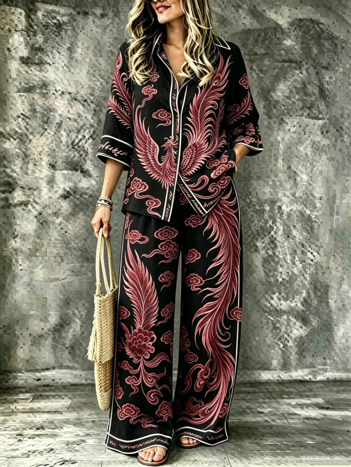 A woman models a matching two-piece pajama set featuring a black base with a large red and pink phoenix and cloud pattern, styled with a woven straw bag against a textured gray wall.