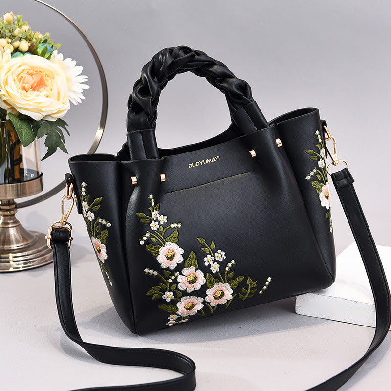 Black handbag with floral embroidery on a white surface with flowers in the background