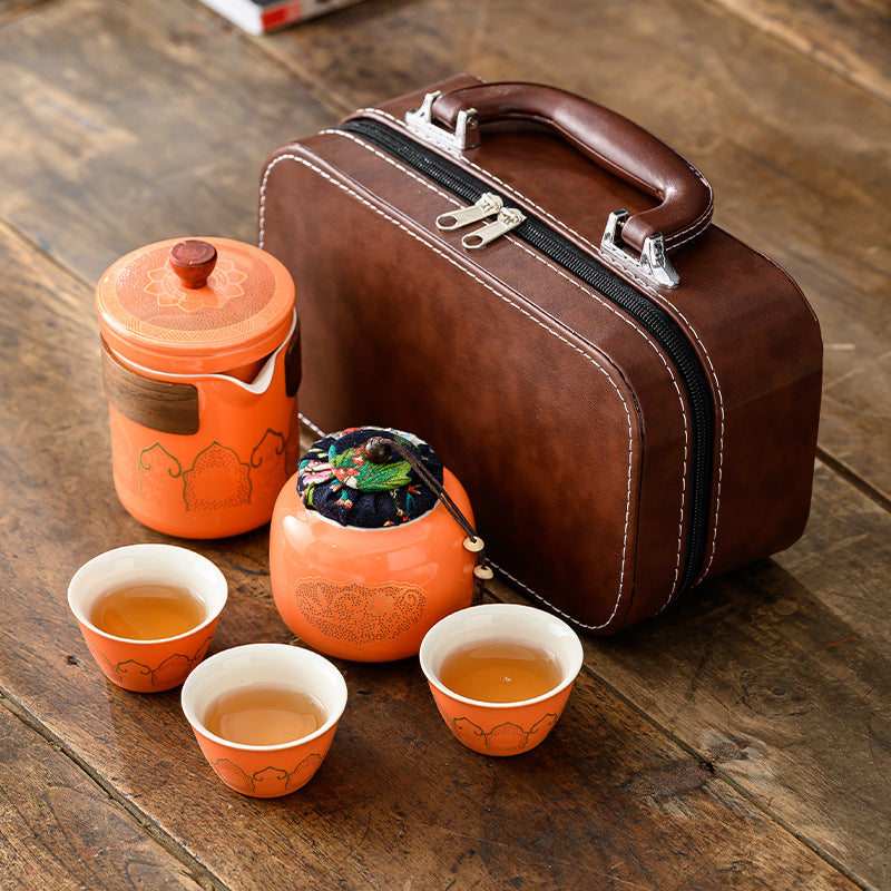 Tea set with orange teapot, cups, and tea leaves on a wooden surface next to a brown leather suitcase.