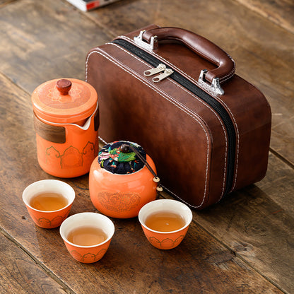Tea set with orange teapot, cups, and tea leaves on a wooden surface next to a brown leather suitcase.