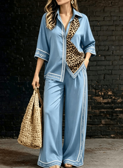 A woman models a light blue two-piece lounge set with white piping and a leopard-print panel detail on the shirt. She is holding a woven straw tote bag against a dark brick wall background.