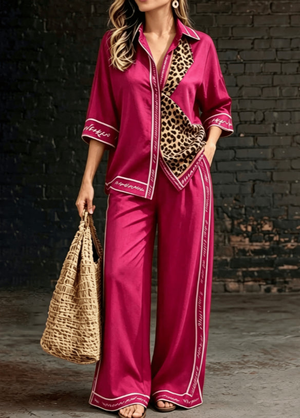 A woman models a two-piece satin pajama set in a deep magenta pink, featuring a button-up shirt with leopard print and white piping, and matching wide-leg pants with white piping. She is holding a woven straw tote bag against a dark brick wall background.