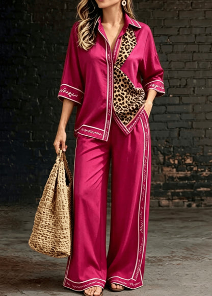A woman models a two-piece satin pajama set in a deep magenta pink, featuring a button-up shirt with leopard print and white piping, and matching wide-leg pants with white piping. She is holding a woven straw tote bag against a dark brick wall background.