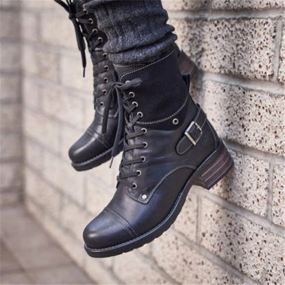 Black leather boots with buckles worn by a person against a brick wall.