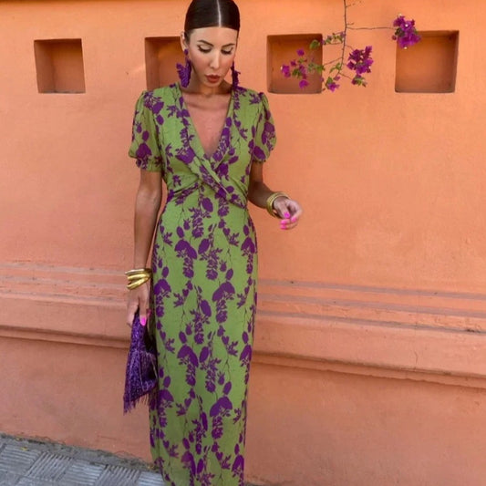 Woman in a green and purple floral dress standing against an orange wall with a plant.