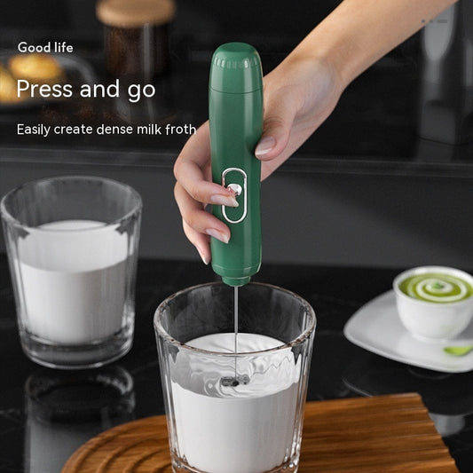 Person using a green handheld milk frother to create froth in a glass on a kitchen counter.