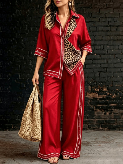 A woman models a red satin pajama set with leopard print panels and white piping details, holding a straw bag against a dark brick wall background.