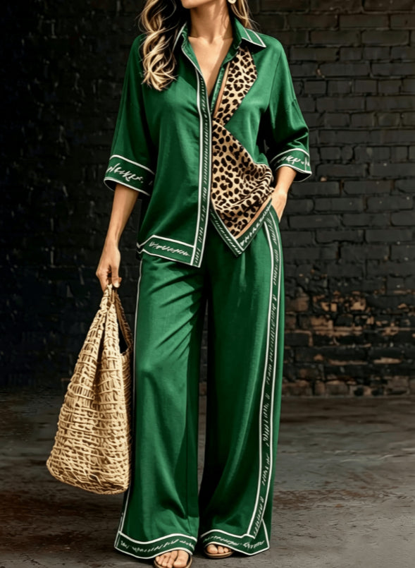 A woman models a green satin pajama set with leopard print lining and white piping details, holding a woven straw bag against a dark brick wall background.