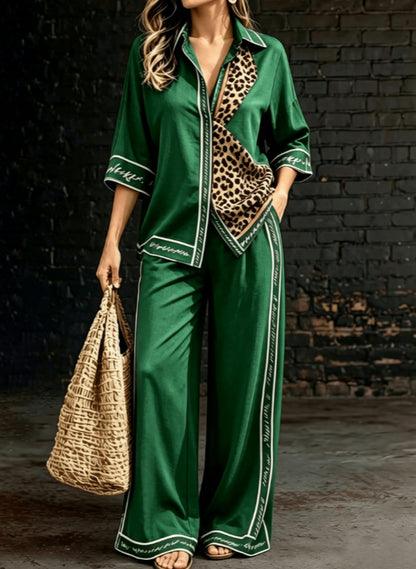 A woman models a green satin pajama set with leopard print lining and white piping details, holding a woven straw bag against a dark brick wall background.
