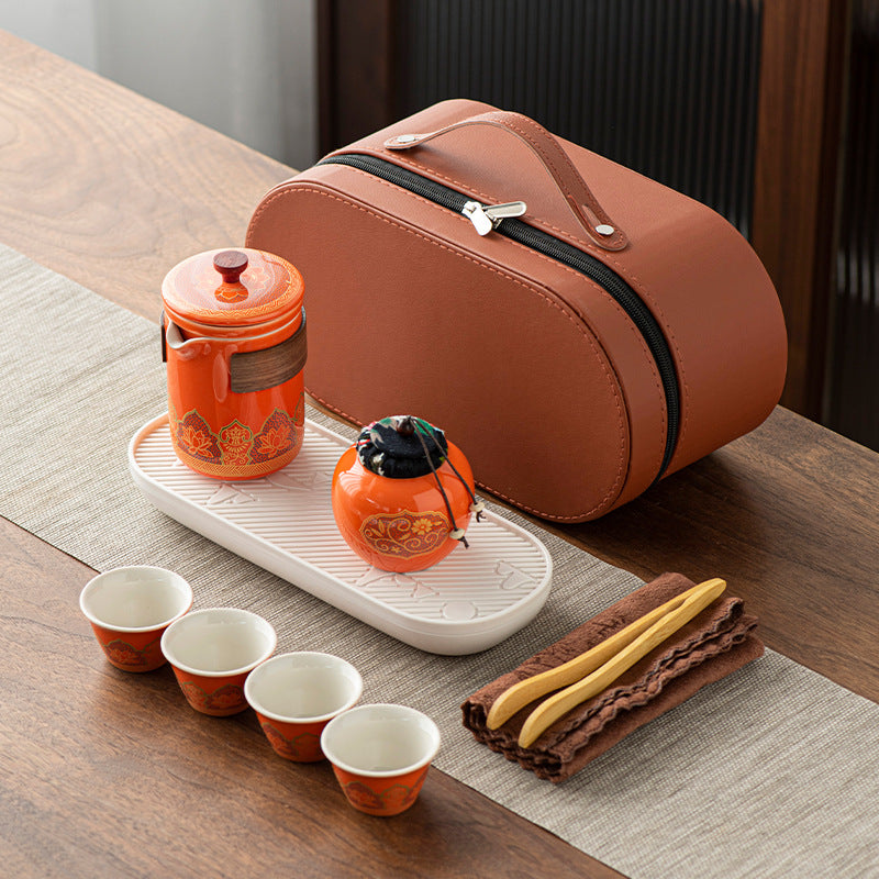 Tea set with orange teapot, cups, and brown carrying case on a wooden table.
