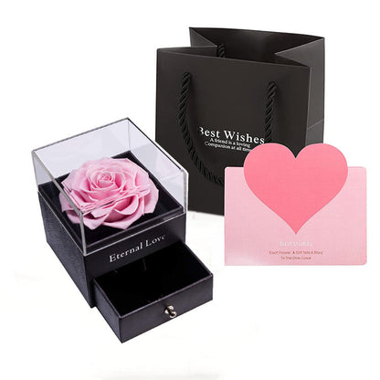 Black box with a pink rose, black gift bag, and pink heart-shaped card on a white background