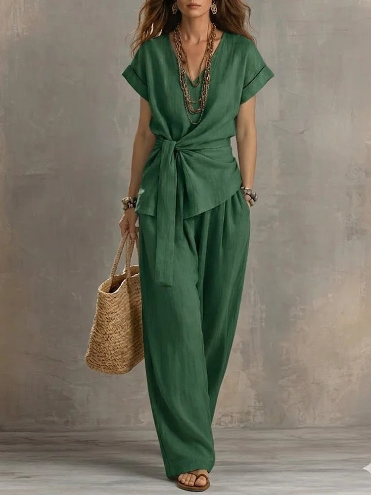 A woman models a green wrap-style short-sleeve jumpsuit with a tie waist and wide-leg pants, holding a straw tote bag against a textured gray studio backdrop.