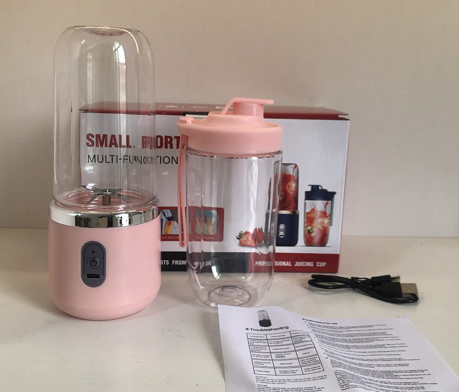 Pink portable blender with clear container, power cable, and user manual on a light surface.