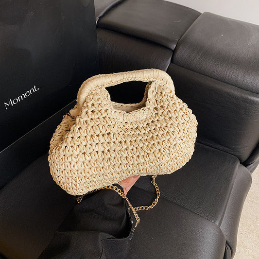 Beige woven handbag with a gold chain held by a person sitting on a black leather couch.