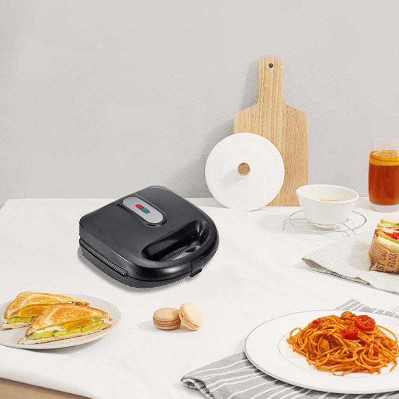 Black sandwich maker on a white table with plates of food and a glass of tea.  YAXSO STORE UAE
