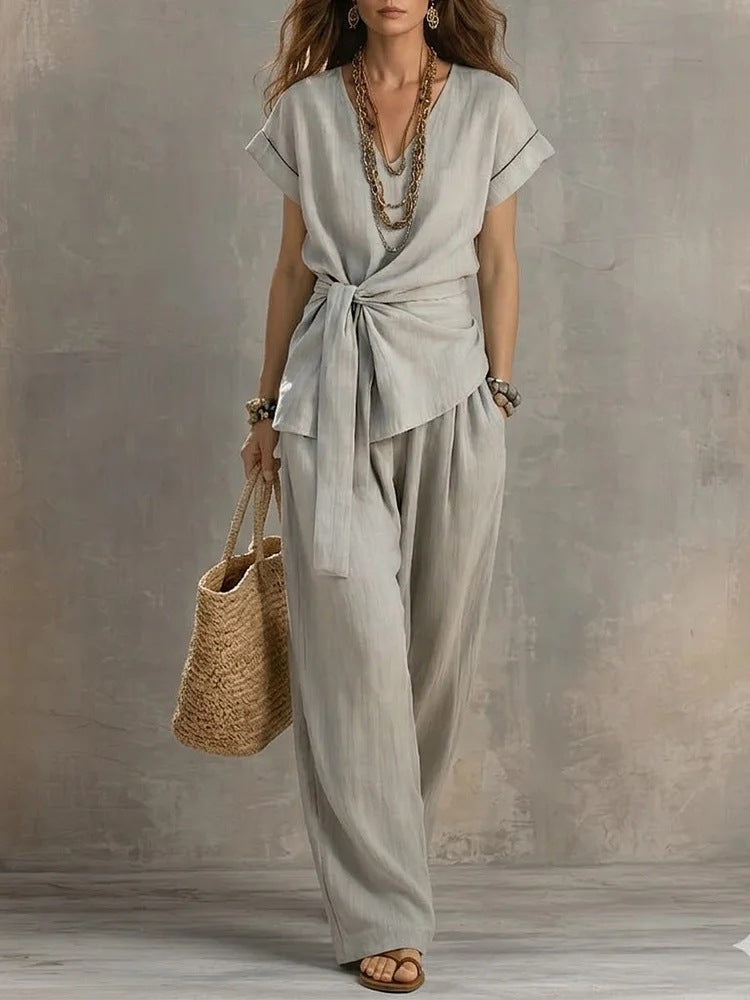 A woman models a light gray linen two-piece set consisting of a short-sleeve wrap top with a tie waist and matching wide-leg pants, styled with layered gold necklaces, bracelets, and a straw tote bag against a textured gray wall.