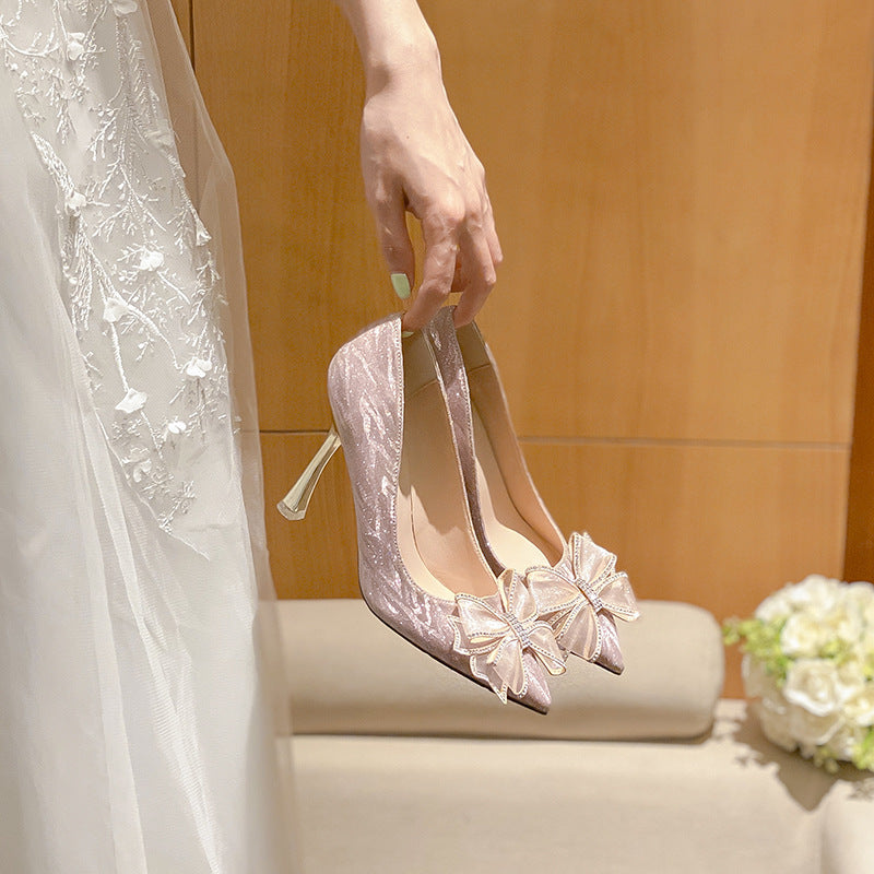 Bridal shoes with decorative bows held by a person wearing a white dress, with a wooden wall and flowers in the background.