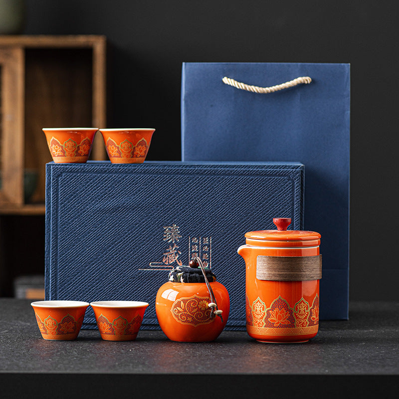 Orange ceramic tea set with four cups and a large container in front of a blue gift box on a dark surface.