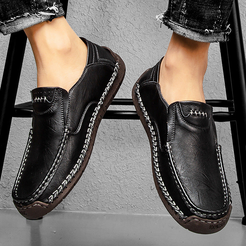 Black leather shoes with white stitching worn by a person sitting on a stool.