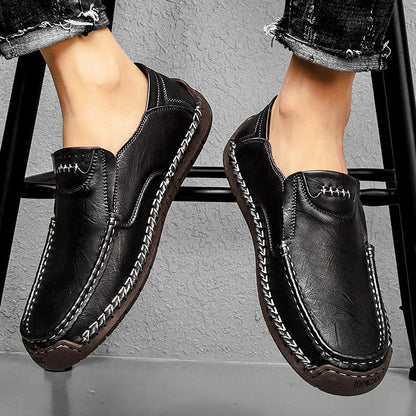 Black leather shoes with white stitching worn by a person sitting on a stool.