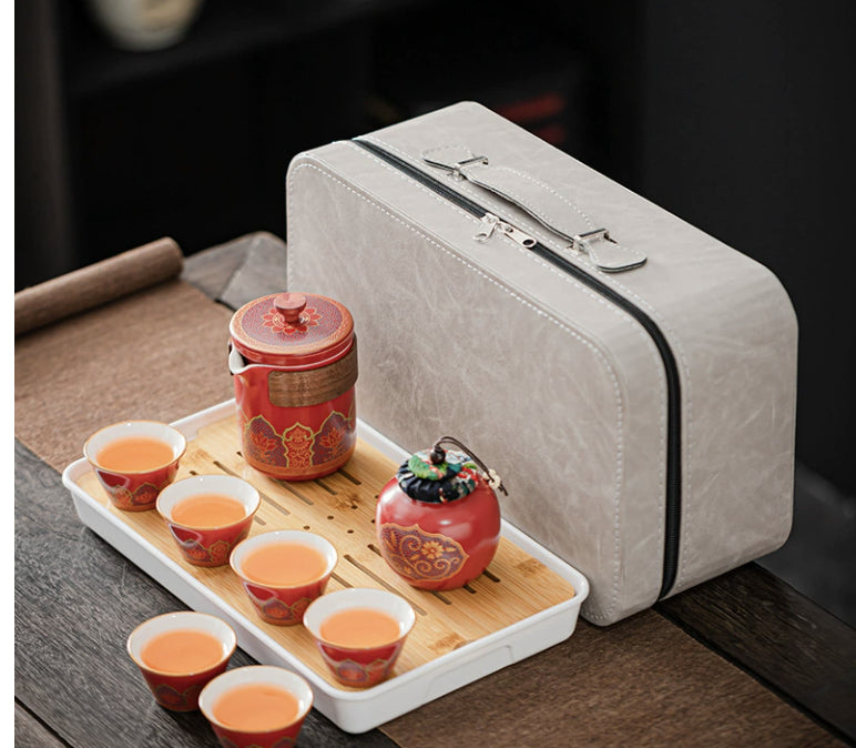 Tea set with red teapot and cups on a wooden tray next to a gray suitcase.