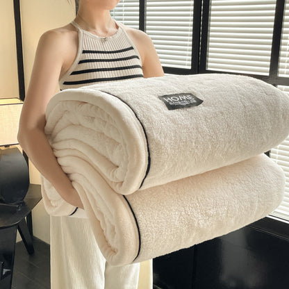 Person holding a folded beige blanket with a brand label in a room with blinds.