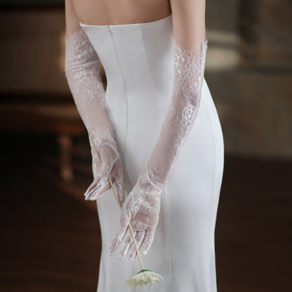Person wearing a white wedding dress with lace gloves holding a flower.