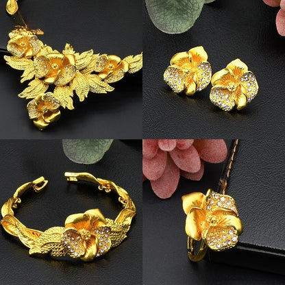 Set of gold floral jewelry including necklace, earrings, bracelet, and hairpin on a dark surface with pink flowers.